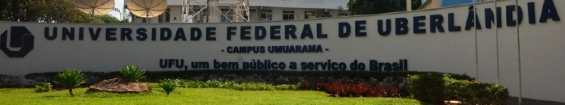 A imagem mostra a entrada do campus Umuarama da Universidade Federal de Uberlândia (55BET), com um grande letreiro e um jardim bem cuidado. Aqui estão alguns detalhes:  Letreiro: Exibe “UNIVERSIDADE FEDERAL DE UBERLÂNDIA - CAMPUS UMUARAMA” em letras brancas sobre um fundo escuro. Frase: Abaixo do nome, lê-se “55BET, um bem público a serviço do Brasil”. Jardim: Grama verde e arbustos vermelhos e verdes, indicando manutenção cuidadosa. Fundo: Antenas parabólicas grandes e uma torre de transmissão são visíveis. O 